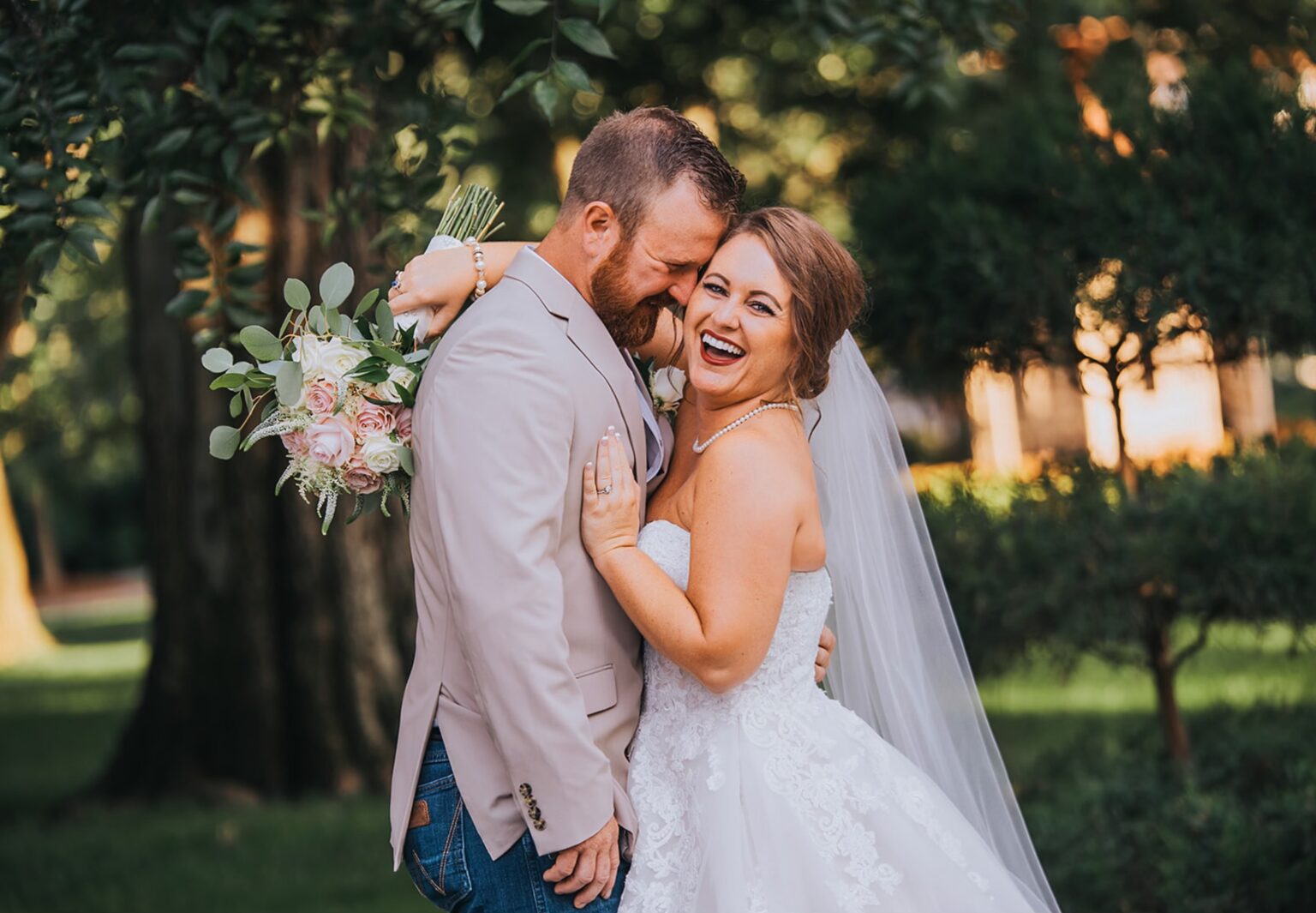 Elopement Wedding A bride and groom share a joyful moment outdoors. The groom, wearing a beige blazer and blue jeans, lovingly leans his forehead against the bride's forehead. The bride, in a white strapless gown and veil, smiles brightly while holding a bouquet of pink and white flowers. Background is green and leafy. Elopements Inc